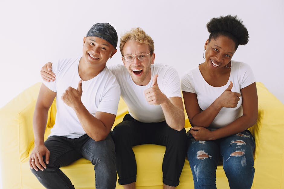 Three happy friends in casual wear giving thumbs up while sitting on a yellow couch.